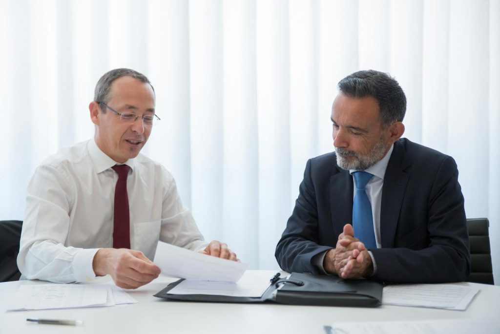 Dos hombres en traje revisando documentos durante una consulta sobre la solicitud de nacionalidad española en una oficina luminosa. Two men in suits reviewing documents during a consultation about Spanish nationality application in a well-lit office.