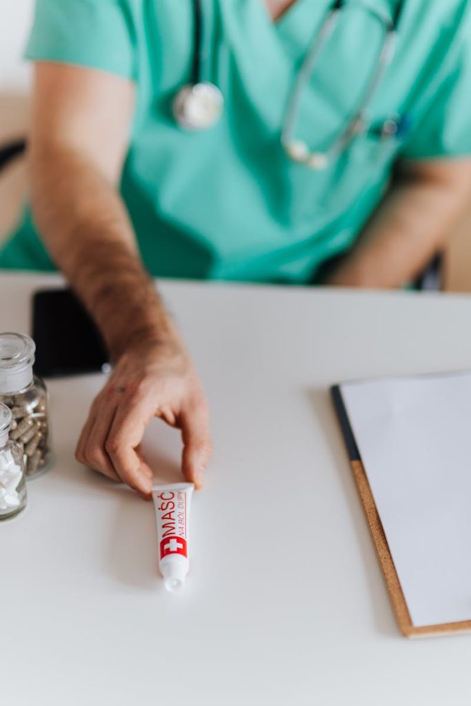 Unrecognizable male doctor wearing medical uniform giving ointment tube to patient while working in modern clinic sitting at table with pills and notebook