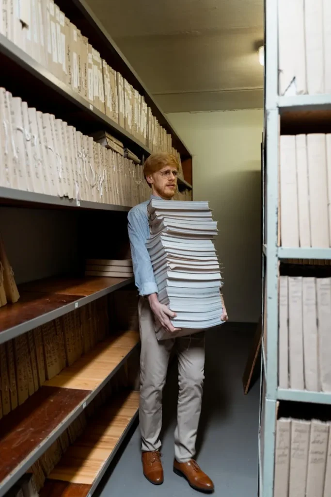 Adult man in archive room carrying a large stack of documents, surrounded by shelves.
