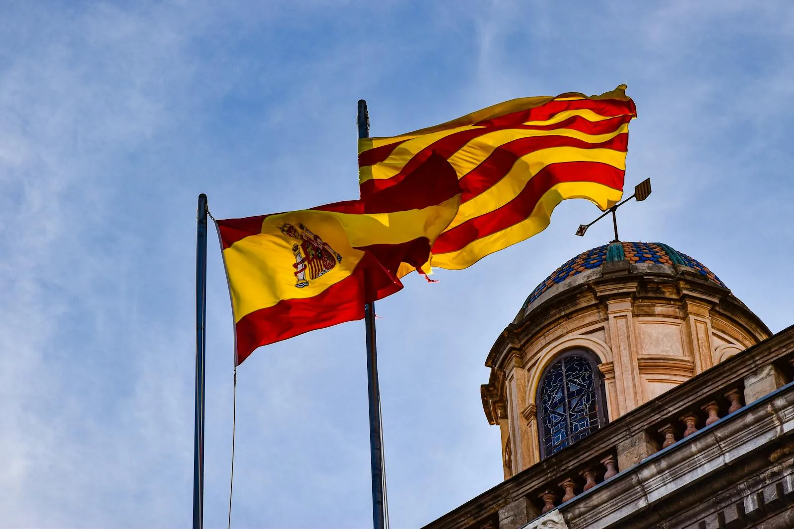 Spanish and Catalan flags waving against a blue sky in front of a historical building.